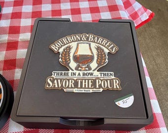 Pode incluir: Um tabuleiro de jogo quadrado preto com um gráfico central apresentando um copo de líquido âmbar, espigas de trigo e o texto "BOURBON & BARRELS THREE IN A ROW... THEN SAVOR THE POUR". O tabuleiro possui uma superfície texturizada.