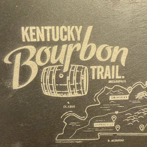Op de afbeelding: Donkergrijze oppervlakte met de woorden "Kentucky Bourbon Trail" in een gestileerd lettertype. Een vat-afbeelding staat tussen de woorden. Een kaart van Kentucky met markeringen staat eronder.