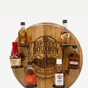 May include: A circular wooden barrel head display rack featuring the engraved text My Kentucky Bourbon Trail. The rack holds six miniature bottles of bourbon, including brands like Maker's Mark, Elijah Craig, Buffalo Trace, and Jim Beam.