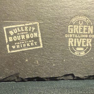 Op de afbeelding: Een donkergrijze leistenen onderzetter met twee witte gedrukte logo's. Het linkerlogo luidt "BULLEIT BOURBON FRONTIER WHISKEY". Het rechterlogo zegt "GREEN RIVER DISTILLING CO." en "EST. 1885".