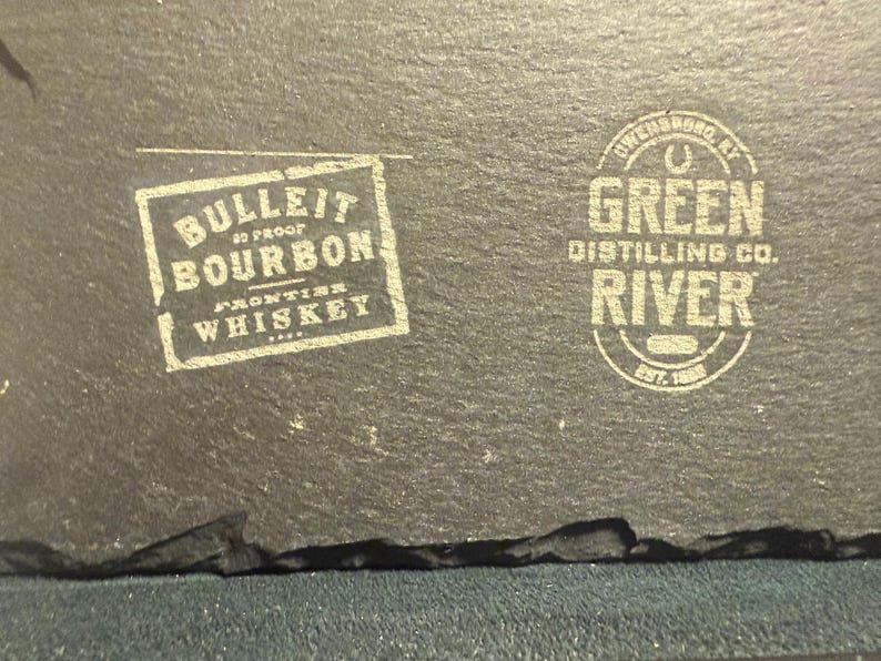 Op de afbeelding: Een donkergrijze leistenen onderzetter met twee witte gedrukte logo's. Het linkerlogo luidt "BULLEIT BOURBON FRONTIER WHISKEY". Het rechterlogo zegt "GREEN RIVER DISTILLING CO." en "EST. 1885".