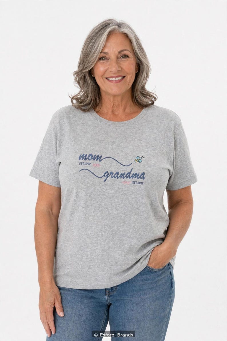 May include: A heather gray short-sleeve t-shirt featuring the text "mom EST.1992" and "grandma EST.2012" in dark blue script, connected by a wavy line with a small yellow and blue bee graphic positioned above the word "grandma."