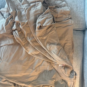 May include: A stack of folded tan t-shirts made of a smooth fabric. The shirts feature a crew neckline and short sleeves, with a visible black graphic print partially showing on the bottom layer of the folded garment stack.