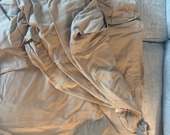 Pode incluir: Uma pilha de camisetas cor de bronze dobradas feitas de tecido liso. As camisetas apresentam gola redonda e mangas curtas, com uma estampa gráfica preta parcialmente visível na camada inferior da pilha de roupas dobradas.