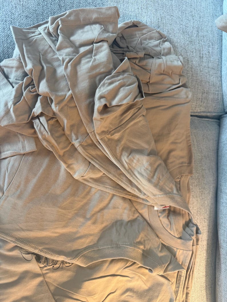 May include: A stack of folded tan t-shirts made of a smooth fabric. The shirts feature a crew neckline and short sleeves, with a visible black graphic print partially showing on the bottom layer of the folded garment stack.