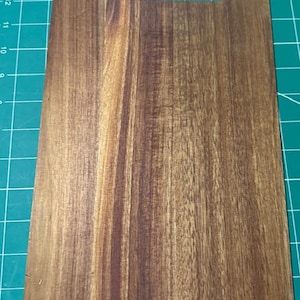 May include: A rectangular wooden cutting board with a smooth, polished finish and a prominent natural grain pattern. The board features a rounded handle cutout at the top center, showcasing rich brown tones and dark wood streaks throughout.