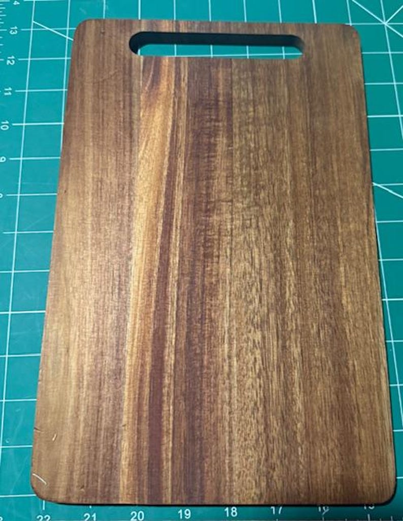 May include: A rectangular wooden cutting board with a smooth, polished finish and a prominent natural grain pattern. The board features a rounded handle cutout at the top center, showcasing rich brown tones and dark wood streaks throughout.