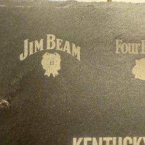 Op de afbeelding: Een donkergrijze leistenen onderzetter met de logo's van Jim Beam en Four Roses in het oppervlak ge&euml;tst. Het Jim Beam-logo is gebogen boven een badge en het Four Roses-logo is een roos.