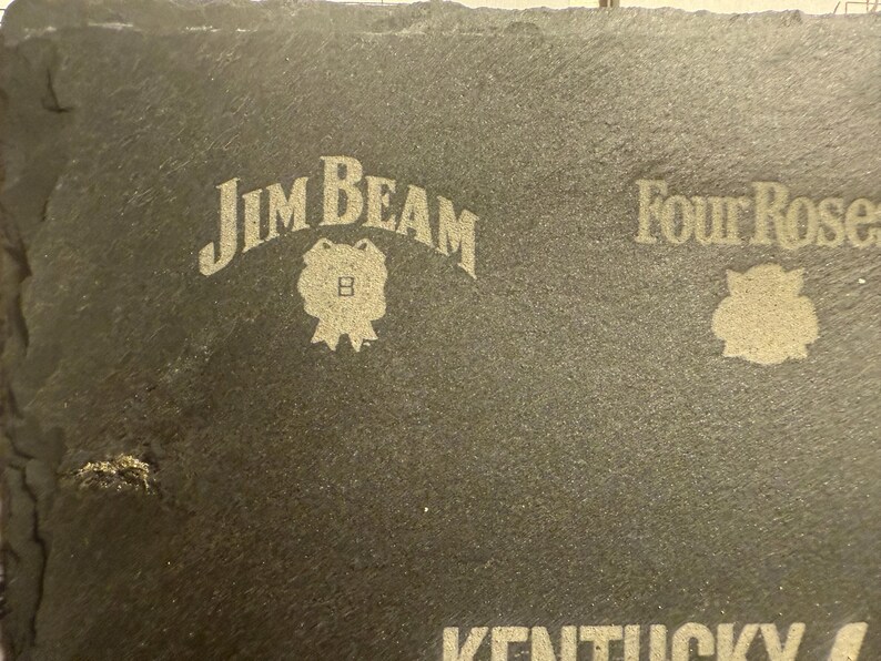 Op de afbeelding: Een donkergrijze leistenen onderzetter met de logo's van Jim Beam en Four Roses in het oppervlak ge&euml;tst. Het Jim Beam-logo is gebogen boven een badge en het Four Roses-logo is een roos.