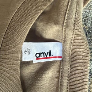 May include: A tan t-shirt with a white interior neck label featuring the brand name anvil in black text with a red horizontal line underneath. The label also includes the text made in uzbekistan and the size L.
