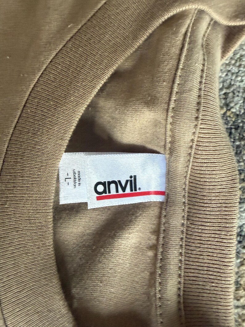 May include: A tan t-shirt with a white interior neck label featuring the brand name anvil in black text with a red horizontal line underneath. The label also includes the text made in uzbekistan and the size L.