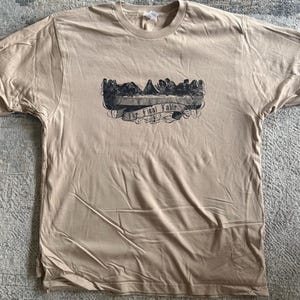 May include: A tan short-sleeve t-shirt featuring a black graphic print on the chest. The design depicts a group of figures seated at a long table with the text The Final Table written on a banner below the illustration in a script font.