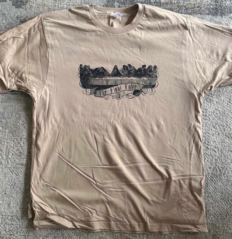 May include: A tan short-sleeve t-shirt featuring a black graphic print on the chest. The design depicts a group of figures seated at a long table with the text The Final Table written on a banner below the illustration in a script font.