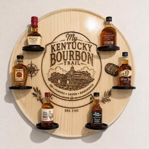 May include: A circular light-coloured wooden wall display featuring the engraved text My Kentucky Bourbon Trail, Est. 1792, Sip, Smoke, Savor, Reminisce. It includes six small black shelves holding miniature bottles of Maker's Mark, Woodford Reserve, Four Roses, Buffalo Trace, Wild Turkey 101, and Evan Williams.