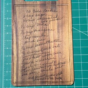 May include: A rectangular wooden cutting board with a handle cutout at the top. The surface is engraved with a handwritten recipe for No Bake Cookies, listing ingredients like sugar, cocoa powder, milk, margarine, vanilla, salt, peanut butter, and oats.