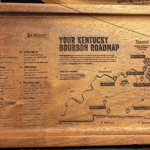 May include: An oval wooden serving tray featuring an engraved map titled Your Kentucky Bourbon Roadmap. The surface includes a detailed list of distilleries and a geographic map of Kentucky with marked locations and text labels.