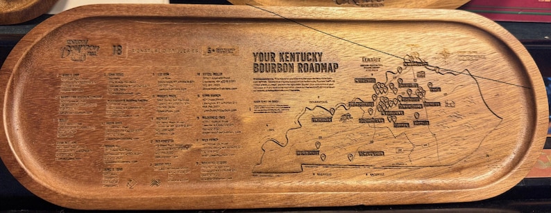 Puede incluir: Una bandeja de servicio ovalada de madera con un mapa grabado titulado Your Kentucky Bourbon Roadmap. La superficie incluye una lista detallada de destiler&iacute;as y un mapa geogr&aacute;fico de Kentucky con ubicaciones marcadas.