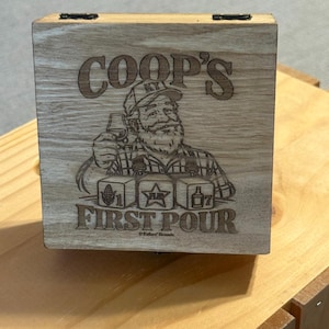 May include: A square wooden box with a hinged lid featuring a laser-engraved illustration of a bearded person wearing a cap, holding a glass. The text reads COOP'S FIRST POUR, with three cubes showing a corn cob, a star, and a bottle.