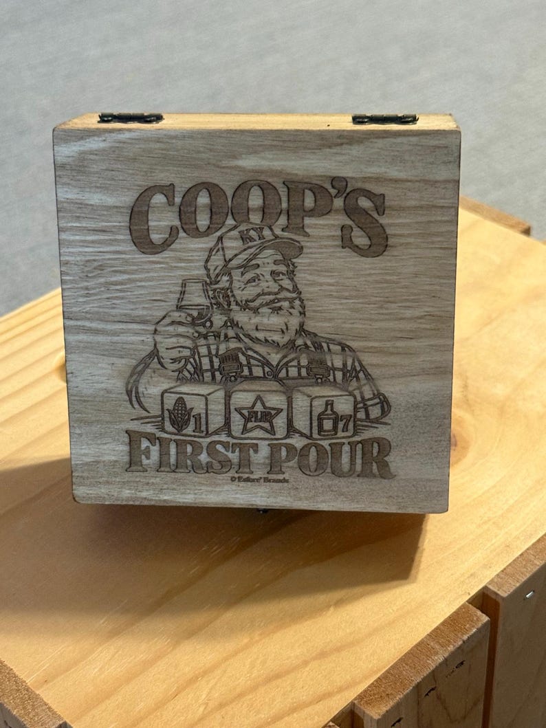 May include: A square wooden box with a hinged lid featuring a laser-engraved illustration of a bearded person wearing a cap, holding a glass. The text reads COOP'S FIRST POUR, with three cubes showing a corn cob, a star, and a bottle.