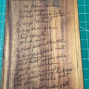 May include: A rectangular wooden cutting board with a handle cutout at the top, featuring a laser-engraved recipe for No Bake Cookies. The text includes ingredients like sugar, cocoa powder, milk, margarine, vanilla, salt, peanut butter, and oats.