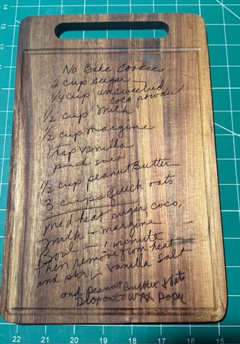 May include: A rectangular wooden cutting board with a handle cutout at the top, featuring a laser-engraved recipe for No Bake Cookies. The text includes ingredients like sugar, cocoa powder, milk, margarine, vanilla, salt, peanut butter, and oats.