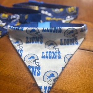 May include: A triangular pet bandana featuring a white background with a repeating pattern of blue Detroit Lions football helmets and the text LIONS in blue lettering. The bandana has a blue fabric trim along the top edge.