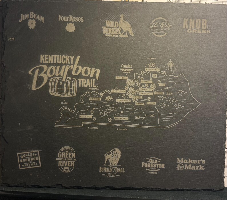 Op de afbeelding: Een donkergrijze leisteen serveerplank met de Kentucky Bourbon Trail kaart en distilleerderij logo's. De plank bevat de tekst "Kentucky Bourbon Trail" en logo's van Jim Beam, Four Roses, Wild Turkey, etc. Een decoratief item.