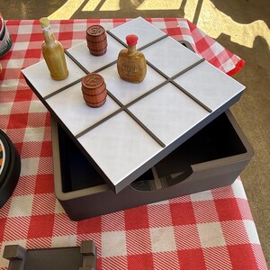 May include: A tic-tac-toe game set featuring a dark brown storage box with a white grid lid. The game pieces include two brown miniature barrels and two small bottles, one yellow and one brown with a red cap, arranged on the grid.