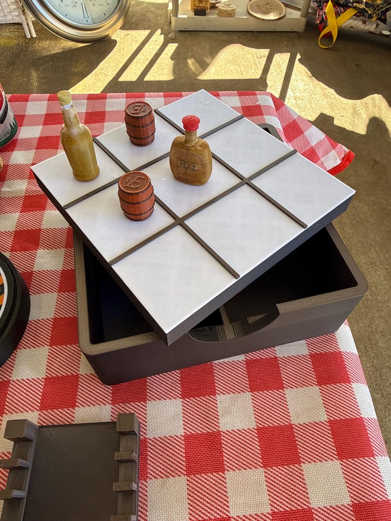 May include: A tic-tac-toe game set featuring a dark brown storage box with a white grid lid. The game pieces include two brown miniature barrels and two small bottles, one yellow and one brown with a red cap, arranged on the grid.