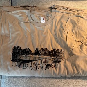 May include: A tan short-sleeved crew neck t-shirt featuring a dark brown graphic print of The Last Supper scene with a banner underneath that reads The Final Table in a stylised font. The shirt has a visible white brand tag at the inner neckline.