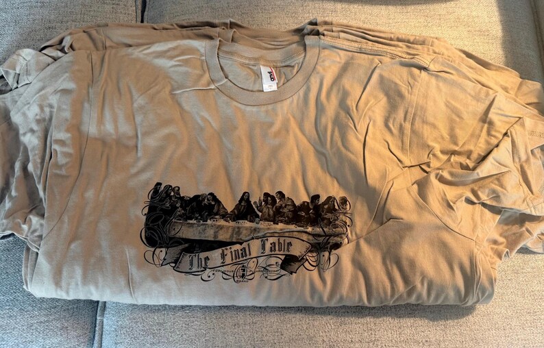 May include: A tan short-sleeved crew neck t-shirt featuring a dark brown graphic print of The Last Supper scene with a banner underneath that reads The Final Table in a stylised font. The shirt has a visible white brand tag at the inner neckline.