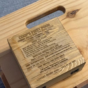 May include: A square wooden box with a light brown grain finish, featuring laser-engraved text on the lid. The text includes the title COOP'S FIRST POUR, instructions on how to play, and rules for the Master Distiller's Die game.