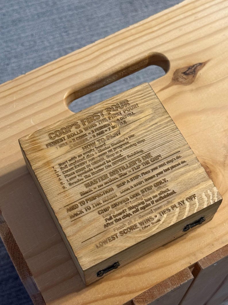 May include: A square wooden box with a light brown grain finish, featuring laser-engraved text on the lid. The text includes the title COOP'S FIRST POUR, instructions on how to play, and rules for the Master Distiller's Die game.