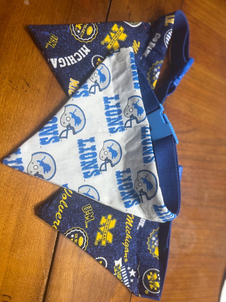 May include: A reversible pet bandana featuring a white fabric side with blue Detroit Lions football helmet graphics and text, and a dark blue fabric side with yellow Michigan Wolverines logos, text, and football icons, secured with a blue plastic buckle.