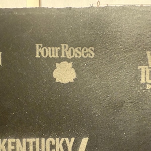 Op de afbeelding: Close-up van een donkergrijze oppervlakte met het "Four Roses" logo en een bloemenembleem in het midden. Andere merknamen, waaronder "Beam" en "Wild Turkey Bourbon", zijn ook zichtbaar.