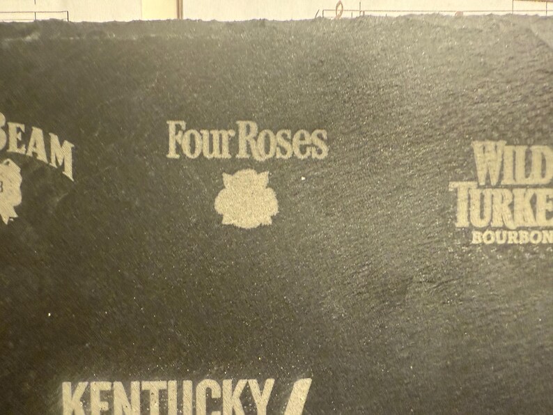 Op de afbeelding: Close-up van een donkergrijze oppervlakte met het "Four Roses" logo en een bloemenembleem in het midden. Andere merknamen, waaronder "Beam" en "Wild Turkey Bourbon", zijn ook zichtbaar.