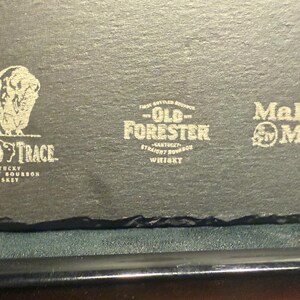 Op de afbeelding: Een donkergrijze leistenen onderzetter met drie whisky-merklogo's: Buffalo Trace, Old Forester en Maker's Mark. Het Buffalo Trace-logo bevat een bizon. De onderzetter heeft een ruwe, natuurlijke rand.