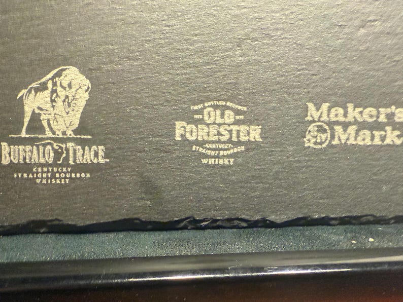 Op de afbeelding: Een donkergrijze leistenen onderzetter met drie whisky-merklogo's: Buffalo Trace, Old Forester en Maker's Mark. Het Buffalo Trace-logo bevat een bizon. De onderzetter heeft een ruwe, natuurlijke rand.