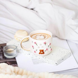 Op de afbeelding: Een witte keramische mok met een gouden handvat, versierd met roze bloemen en een bruine beer. De mok is gevuld met een cappuccino en staat op een wit gevlekt oppervlak.