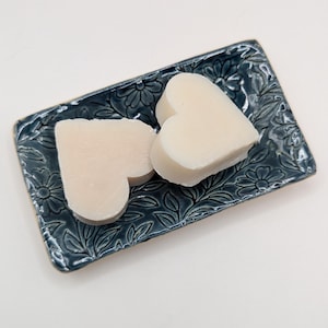 May include: Two heart-shaped, off-white soaps sit on a rectangular, dark teal soap dish. The dish has a floral and leaf pattern. The soaps are smooth and the dish has a glossy finish.