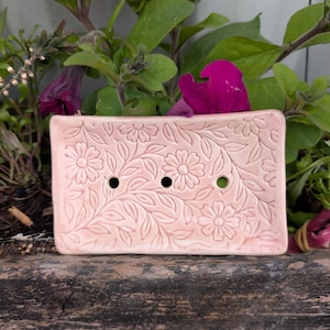 May include: A rectangular, pale pink ceramic soap dish with a floral and leaf design. The dish has three drainage holes. It is set against a backdrop of green leaves and purple flowers.