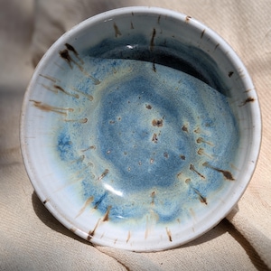 May include: A round ceramic bowl featuring a white rim and a blue glazed interior. The centre of the bowl displays a mottled blue and light yellow finish, with brown drip-like streaks extending from the rim towards the centre of the basin.
