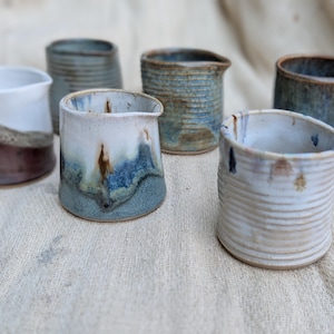 May include: A collection of six ceramic creamers featuring various glazes and textures. The vessels display a range of finishes including smooth white, deep blue, earthy brown, and ribbed patterns, each with a small pouring spout and open top.