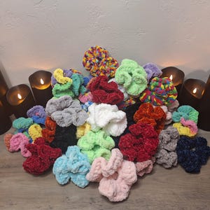 May include: A collection of colorful, handmade scrunchies in various shades of red, blue, green, pink, yellow, and gray. The scrunchies are made of a soft, plush material. Lit candles in dark brown holders are in the background.