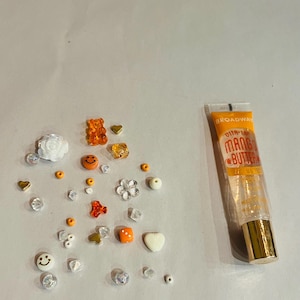 May include: A collection of small, colorful beads and charms, including a white rose, a gummy bear, a smiley face, a flower, a heart, and a dice. A tube of clear lip gloss with the text "Ultra-Shine Mango Butter" and "Broadway" is also included.