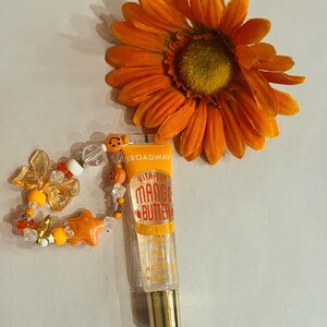 May include: A clear lip gloss tube with a gold cap and a yellow and orange label that says "Vita-Lip Mango Butter Lip Gloss". The tube is attached to a colorful beaded charm bracelet with a star, butterfly, and smiley face charms.
