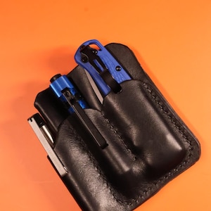 Könnte beinhalten: Ein schwarzes Lederetui mit drei Fächern zum Aufbewahren eines blauen Klappmessers, eines silbernen Stifts und eines schwarzen Stifts.