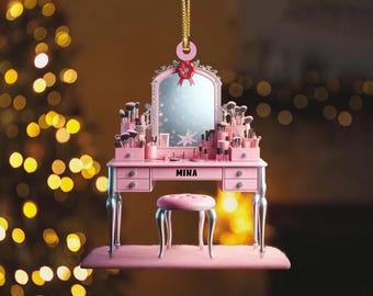 May include: A pink vanity ornament with a mirror, drawers, and a stool. The vanity is adorned with makeup brushes and products. The name "MINA" is displayed on the front. A red bow and gold hanging string complete the design.