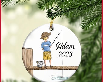May include: A white, round Christmas ornament with a drawing of a person fishing on a pier. The person is wearing a hat, blue shirt, and yellow shorts. The ornament has the name "Adam" and the year "2023" printed on it. A gold string is attached for hanging.