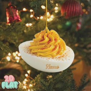 May include: A deviled egg Christmas ornament with a yellow filling and a dusting of paprika. The white egg-shaped ornament has the word "Name" in a cursive font. The ornament hangs from a gold string.